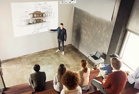 a professor in front of a classroom pointing at a projected image of an architectural drawing of a building