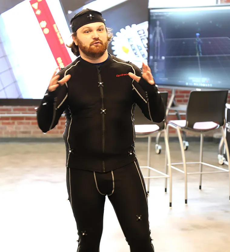 a man motions unthusiastically to a classroom while wearing a full motion capture suit.