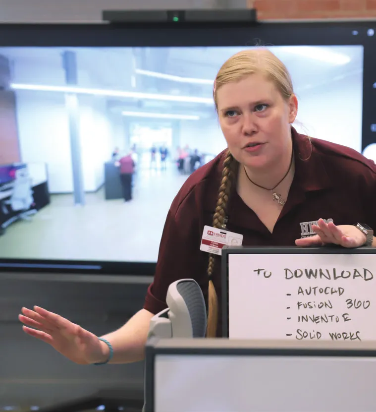 an instructor addresses the class as she holds a whiteboard where she has written a list of 3d modeling software headed with the text 'to download'.  the class itself is visible on the viewboard in the background through a webcam mounted to the unit.