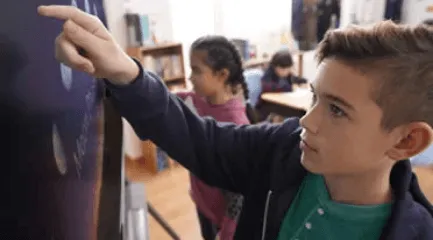 Boy standing in front of and using a ViewSonic ViewBoard display in a classroom with other students.